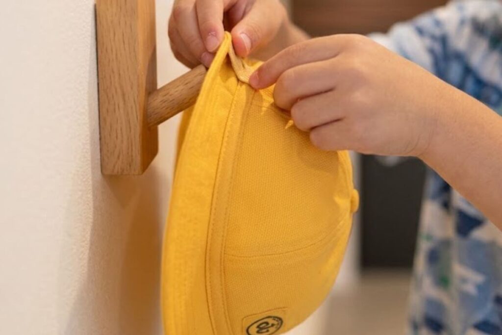 A child's hands carefully hanging a yellow school hat onto a wooden wall-mounted hook.