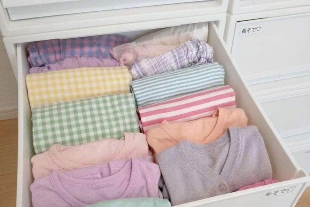 A neat Japanese wardrobe organization setup showing colorful kids' clothes folded vertically inside solid white MUJI storage drawers with a neat Japanese label maker tag.
